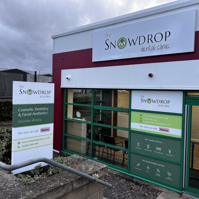 a photo of a wide view of all the signage for snowdrop clinic. Features the double sided totem standing by the walkway, giving passers-by more information. Also there is the new main facia of the store - a white aluminium composite tray with push through letters with a black face and a white glowing halo illumination, reading "the Snowdrop Dental Clinic".